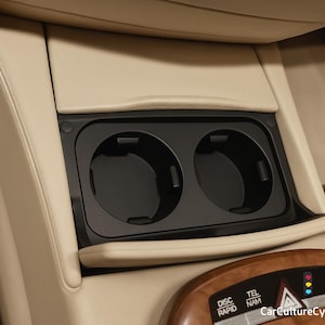 Può includere: Un portabicchieri nero integrato per auto con due scomparti circolari. Il portabicchieri è incastonato in un pannello interno beige, con un cruscotto in legno visibile in basso. L'interno dell'auto ha un aspetto lussuoso.