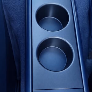 Puede incluir: Primer plano del interior azul de un coche con un portavasos doble. El portavasos está integrado en la consola central, con dos aberturas circulares. El diseño es elegante y moderno, con un acabado mate.