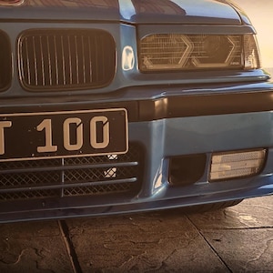 May include: A blue car front end featuring a black grille, rectangular headlights with integrated circular lenses, and a black license plate displaying the text NT 100. The bumper includes a lower mesh grille and a clear rectangular fog light.
