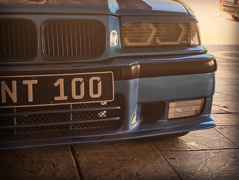 May include: A blue car front end featuring a black grille, rectangular headlights with integrated circular lenses, and a black license plate displaying the text NT 100. The bumper includes a lower mesh grille and a clear rectangular fog light.