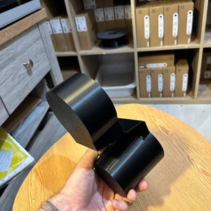 May include: A black, 3D-printed object featuring a cylindrical base connected to a larger, rounded, flat-topped component. The item has a smooth, matte finish and a geometric, multi-part structure designed with distinct, solid, dark-colored segments.