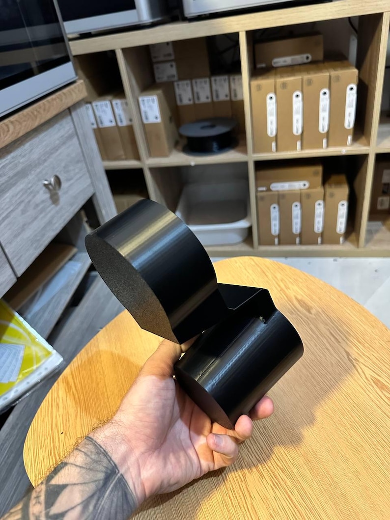 May include: A black, 3D-printed object featuring a cylindrical base connected to a larger, rounded, flat-topped component. The item has a smooth, matte finish and a geometric, multi-part structure designed with distinct, solid, dark-colored segments.
