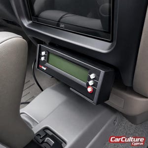 May include: A black ScanGauge automotive diagnostic computer mounted in a vehicle interior. The device features a rectangular green digital display screen, two white buttons on the left side, and two white buttons with one red button on the right side.