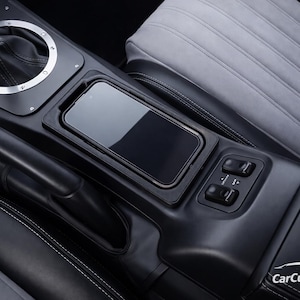 May include: A black automotive centre console featuring a gear shifter with a silver trim ring, a rectangular smartphone storage slot, and two window control switches. The console is positioned between two grey and black upholstered car seats.
