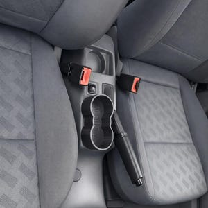 Puede incluir: Portavasos negro para coche y accesorio de aspiradora. El portavasos está diseñado para encajar en la consola central de un coche. Los asientos son grises con un diseño estampado. Los cinturones de seguridad tienen botones de liberación rojos.