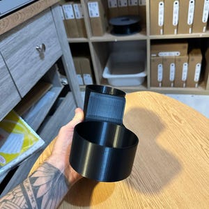 May include: A black 3D-printed cup holder attachment featuring a cylindrical cup-shaped vessel connected to a vertical mounting bracket with a flat rectangular back panel, designed for secure placement on a flat surface or edge.