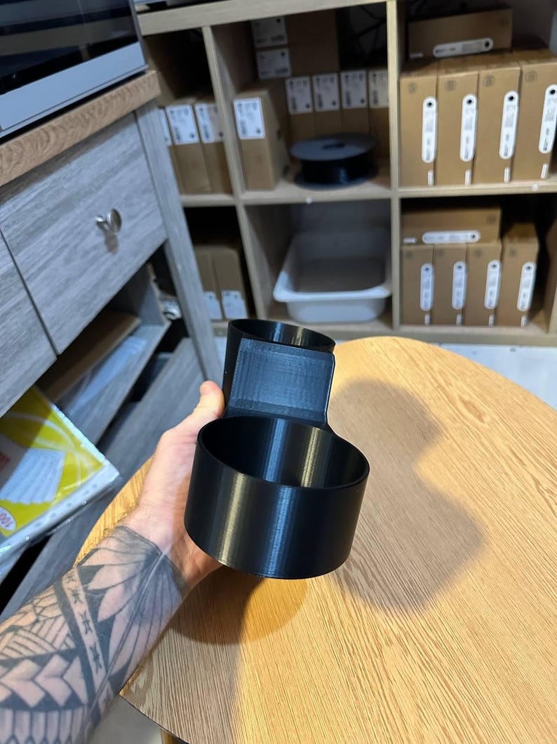 May include: A black 3D-printed cup holder attachment featuring a cylindrical cup-shaped vessel connected to a vertical mounting bracket with a flat rectangular back panel, designed for secure placement on a flat surface or edge.