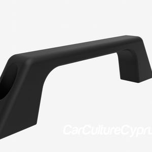 Puede incluir: Un asa negra arqueada con una parte superior rectangular y soportes angulados. El asa tiene dos aberturas circulares en un lado. El producto es probablemente para un vehículo o una aplicación similar. El texto "CarCultureCyprus" es visible.