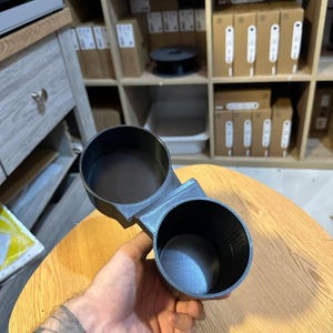 May include: A black dual cup holder accessory featuring two circular openings connected by a central bridge. The object has a textured finish and is constructed with a rigid, hollow design, providing two distinct compartments for holding beverage containers.
