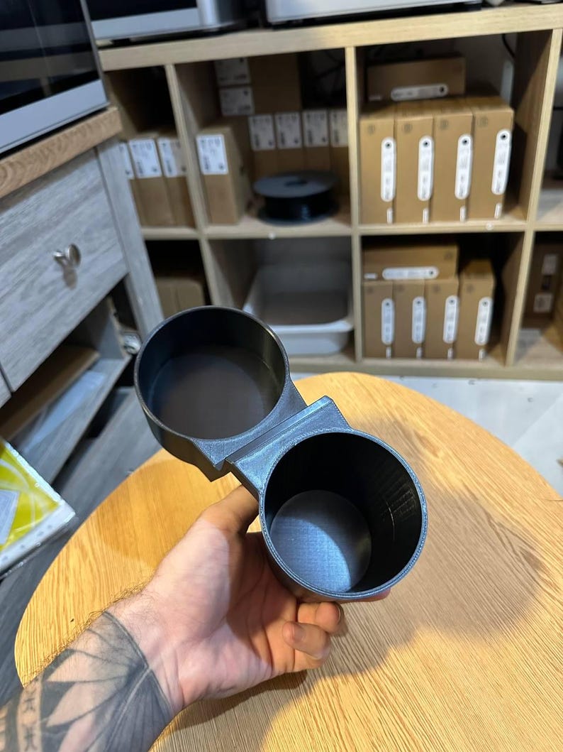 May include: A black dual cup holder accessory featuring two circular openings connected by a central bridge. The object has a textured finish and is constructed with a rigid, hollow design, providing two distinct compartments for holding beverage containers.