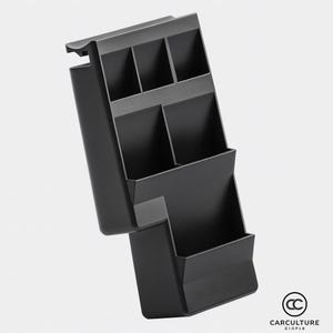 May include: A black car organiser with multiple compartments. The organiser has a stepped design with open storage spaces of varying sizes. The top section has four small rectangular slots, while the lower section has two larger compartments. The organiser is made of a matte black material.