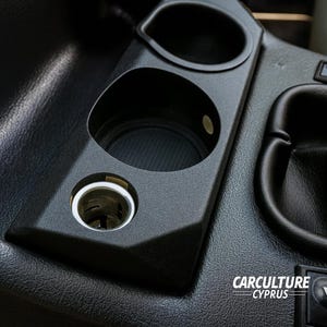 Puede incluir: Consola interior de coche negra con un portavasos incorporado, un pequeño compartimento de almacenamiento y un puerto para encendedor de cigarrillos. La consola es de plástico negro texturizado.