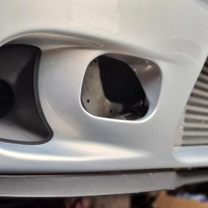 May include: A close-up view of a silver car bumper, showcasing three openings. The bumper has a circular vent on the left, a rectangular one in the middle, and a ribbed section on the right. The car's design is streamlined and contemporary.
