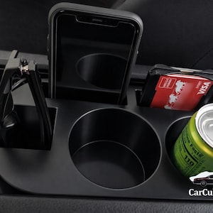 May include: Black car console organizer with multiple compartments. Features a phone holder, cup holders, and a slot for a credit card. Includes a can of soda and a pair of glasses. The organizer is designed to fit in a car's center console.