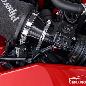 Può includere: Primo piano di un motore di auto con un filtro dell'aria nero con l'etichetta "Pipercross". Il motore include un tubo di aspirazione nero, morsetti metallici e cavi, su sfondo rosso.