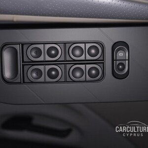 Puede incluir: Primer plano de un panel de control interior de coche negro. El panel presenta una serie de botones redondos, botones direccionales y un solo botón rectangular. La superficie es negro mate con una textura.