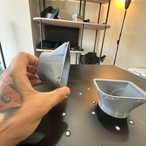 May include: Two grey, 3D-printed plastic air vent adapters with a tapered, funnel-like shape. One adapter is held in a hand, showing its side profile, while the other sits on a flat surface. Both feature a wide, square-shaped opening.