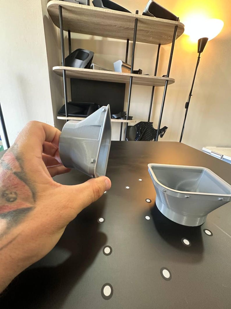 May include: Two grey, 3D-printed plastic air vent adapters with a tapered, funnel-like shape. One adapter is held in a hand, showing its side profile, while the other sits on a flat surface. Both feature a wide, square-shaped opening.
