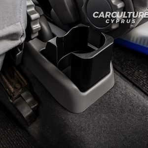 May include: A black car cup holder with a gray trim, installed in a vehicle. The text "CARCULTURE CYPRUS" is visible on the right side of the image. The cup holder is designed to fit into the car's interior.