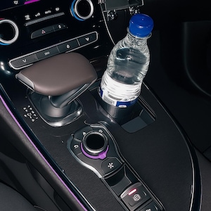 Op de afbeelding: Een doorzichtige plastic waterfles met een blauwe dop staat in de bekerhouder van een auto. Het interieur van de auto heeft een donker dashboard met een bruine versnellingspook en een bedieningspaneel met paarse accenten.
