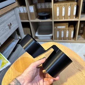 May include: A black plastic dual cup holder accessory featuring two cylindrical compartments connected by a central bridge piece. The object has a smooth, matte finish and is designed to hold two beverage containers simultaneously in a side-by-side configuration.