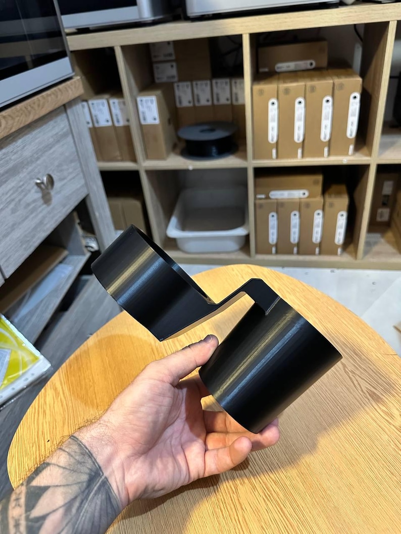 May include: A black plastic dual cup holder accessory featuring two cylindrical compartments connected by a central bridge piece. The object has a smooth, matte finish and is designed to hold two beverage containers simultaneously in a side-by-side configuration.