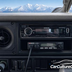 May include: Vehicle dashboard with a Kenwood stereo and a smaller radio unit. The black dashboard includes an air vent and other controls. Snow-capped mountains are visible through the windscreen in the background.