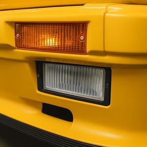 Op de afbeelding: Close-up van een felgele autobumper met een rechthoekige amberkleurige richtingaanwijzer en een rechthoekige koplamp met een zwart frame. De bumper heeft een zwarte onderlip.