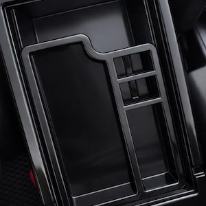 May include: A black car console organizer with multiple compartments. The organizer has a glossy finish and is designed to fit inside a vehicle's center console. It includes a section with dividers and a larger open area.