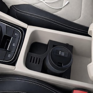 Può includere: Dettaglio degli interni dell'auto con una tazza da viaggio nera in un portabicchieri della console centrale. La console è nera e beige con un cavo di ricarica bianco. I sedili sono grigi e neri con cuciture bianche.