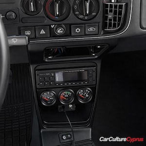 Puede incluir: Panel de instrumentos negro de coche con radio, tres indicadores y varios botones de control. Los indicadores tienen agujas rojas y marcas blancas. La radio tiene una pantalla digital. Interior del coche.