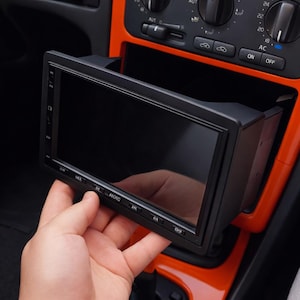 Può includere: Un'unità multimediale per auto rettangolare nera con display touchscreen e pulsanti fisici lungo il bordo inferiore, con il marchio ATOTO stampato sulla parte anteriore, posizionata in un cruscotto con finiture arancioni brillanti.