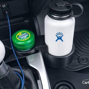 May include: A black plastic car cup holder expander installed in a vehicle center console. It holds a white water bottle with a blue logo and a small green circular container labeled Excitemint. A white gear shift knob is visible in the foreground.
