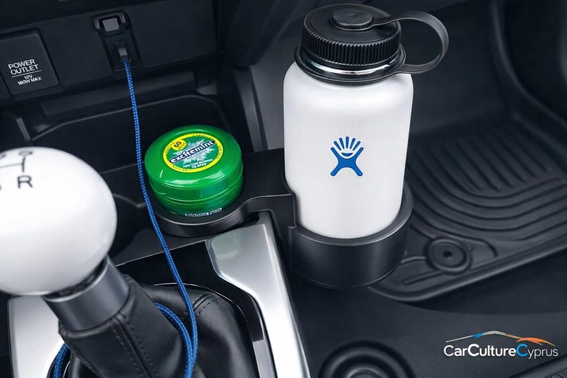 May include: A black plastic car cup holder expander installed in a vehicle center console. It holds a white water bottle with a blue logo and a small green circular container labeled Excitemint. A white gear shift knob is visible in the foreground.
