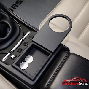 May include: Black car interior with a cup holder and storage compartment. The cup holder is attached to the console and has a circular opening. Two silver coins are in the storage area. The interior is black and beige.