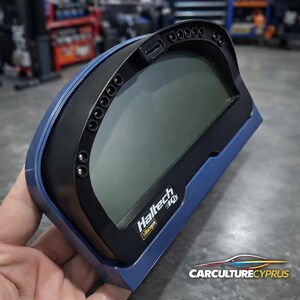 May include: A blue and black digital dashboard with the brand name "Haltech" visible. The dashboard has a rectangular screen and several small circular buttons along the top edge. The device is held in a hand.
