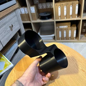 May include: A black plastic dual cup holder accessory featuring two cylindrical compartments connected by a central bridge. The object has a smooth, matte finish and a rigid structure designed to hold two beverage containers simultaneously.