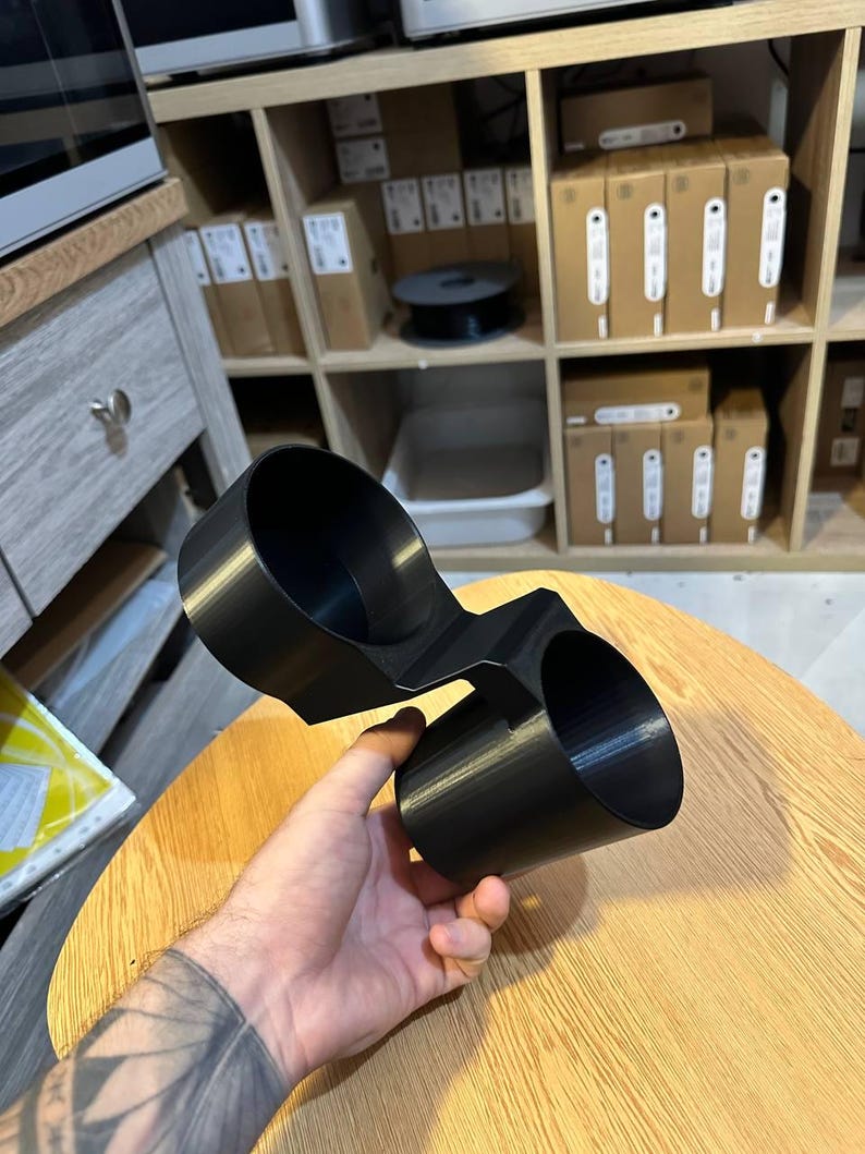 May include: A black plastic dual cup holder accessory featuring two cylindrical compartments connected by a central bridge. The object has a smooth, matte finish and a rigid structure designed to hold two beverage containers simultaneously.