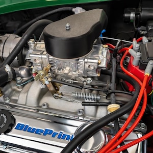 Puede incluir: Primer plano de un motor de coche clásico. El motor presenta un filtro de aire negro, un carburador plateado y cables de bujías rojos. El bloque del motor es cromado con la palabra "BluePrint" en letras azules. El fondo es verde.