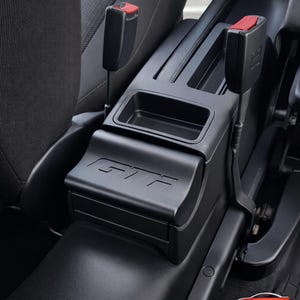 Könnte beinhalten: Schwarze Autokonsole mit den Buchstaben "GTF" auf der Vorderseite. Die Konsole hat ein Staufach und befindet sich zwischen zwei Sicherheitsgurtschnallen mit roten Entriegelungsknöpfen. Innenraum eines Autos.