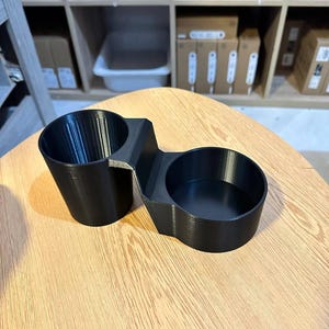 May include: A black 3D-printed dual cup holder featuring two circular compartments of different depths connected by a central bridge. The object has a smooth, matte finish with visible horizontal layer lines from the manufacturing process.