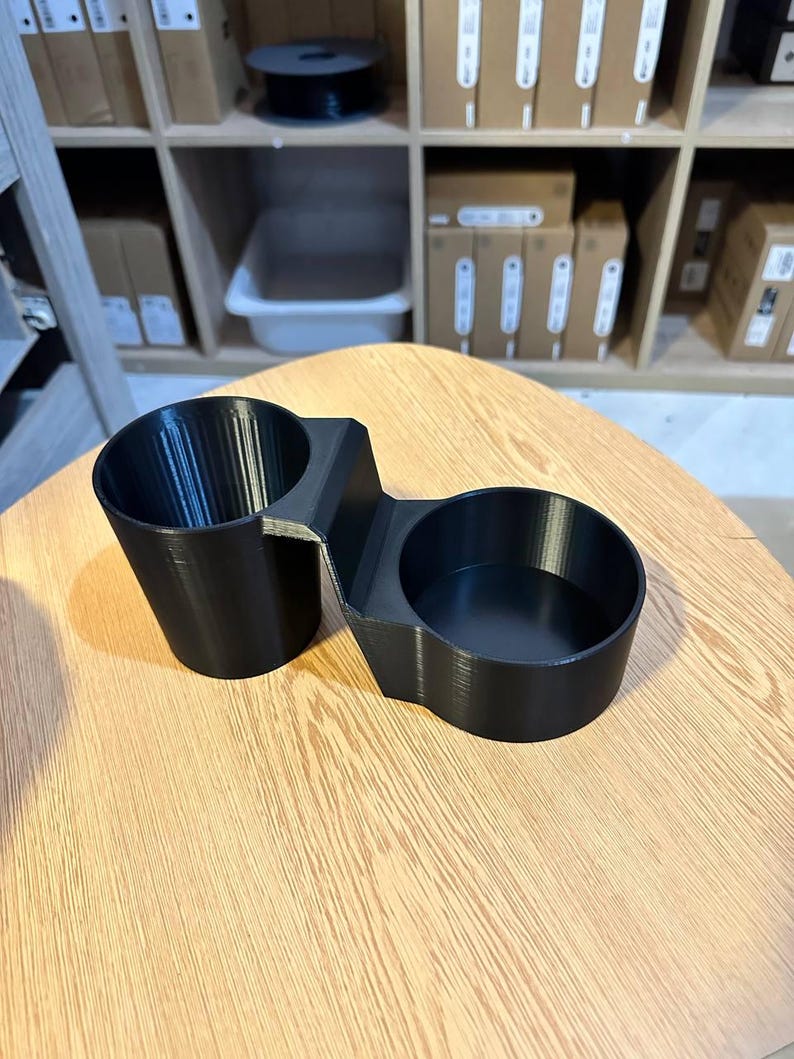 May include: A black 3D-printed dual cup holder featuring two circular compartments of different depths connected by a central bridge. The object has a smooth, matte finish with visible horizontal layer lines from the manufacturing process.