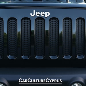May include: Close-up of a black Jeep Wrangler's front grille. The word "Jeep" is displayed in white above the grille. The grille has vertical slots with a textured pattern. Headlights are visible on either side.