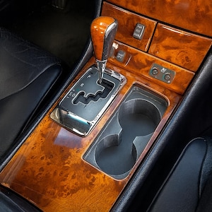 Op de afbeelding: Auto-interieurconsole met een houtnerf afwerking. De console heeft een versnellingspook, bekerhouders en bedieningsknoppen. De houtnerf heeft een warme, roodachtig bruine tint, en de console staat tegen een zwart interieur.