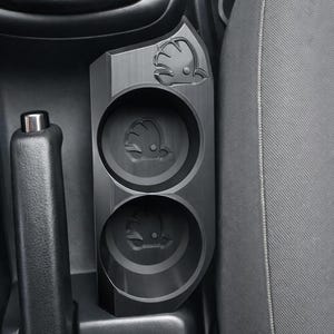 Puede incluir: Un portavasos negro para coche con dos aberturas circulares, con el logotipo de Skoda. El soporte está situado entre los asientos del coche y la consola central. El diseño es elegante y moderno, con un acabado de metal cepillado.