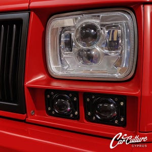 May include: A close-up of a red car's front, featuring a black grill and a large, square headlight. Beneath the headlight are two smaller, round lights. The image includes the "Car Culture Cyprus" logo in the bottom right.