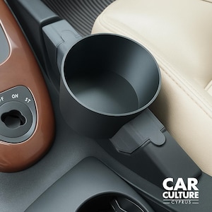 May include: A matte black plastic car cup holder insert with a cylindrical shape, designed to fit into a vehicle center console. The accessory features a smooth interior and a textured exterior, positioned next to a brown control panel with the text ON ST.
