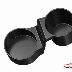 May include: A black dual cup holder with two circular openings connected by a central bridge. The object features a smooth, matte finish and a solid construction. In the bottom right corner, the text CarCultureCyprus appears in black and red.