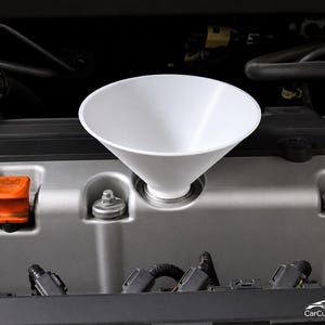 May include: A white plastic funnel inserted into the oil filler port of a silver engine valve cover. An orange oil cap is visible to the left, alongside various black rubber hoses and metal bolts within the engine bay of a vehicle.