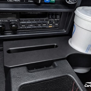 Puede incluir: Consola de coche negra con un portavasos incorporado y una ranura de almacenamiento rectangular. Un vaso de papel blanco con una pajita azul está en el portavasos. El sistema de sonido del coche es visible en el fondo.